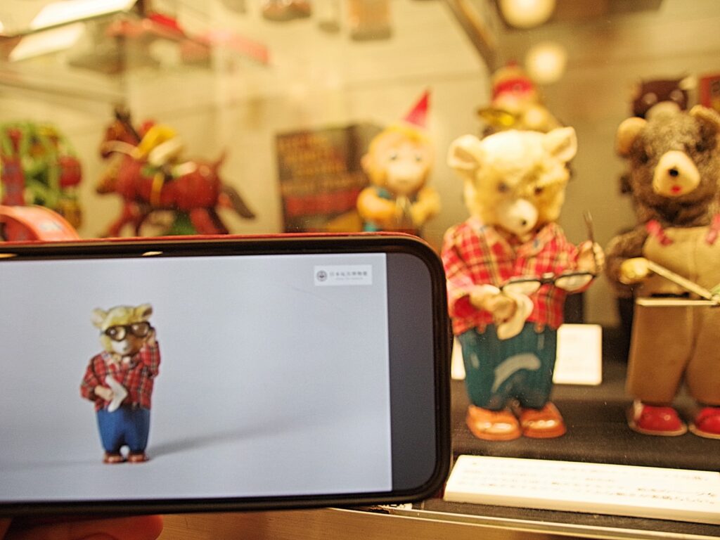 いくつかの資料の説明書きに二次元コードがあり、スマホで読み取ると動画を閲覧できる。写真は、ゼンマイ仕掛けの「メガネをふくクマ」が動く様子のひとこま